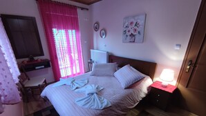 A bedroom with a bed, a bedside table with a lamp, a window with pink curtains, a wall-mounted artwork, and a checkered floor. - Αρχοντικό Δρυάδες 1 (Belokomiti)
