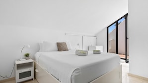 A modern bedroom with a large bed, a bedside table with a lamp, and a view of the outside through a sliding door.