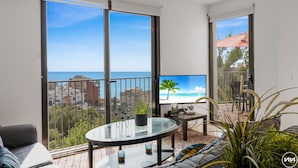 A balcony with a glass table, a sofa, and a TV displaying a beach scene.