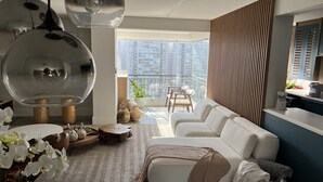 Una sala de estar moderna con un sofá blanco, una mesa de centro y un comedor con mesa y sillas blancas. Hay un balcón con vistas a la ciudad.