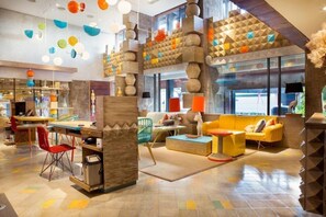 Relax in our vibrant lobby, featuring a central seating area with a sofa, ottoman, and chairs, surrounded by colorful geometric patterns and unique lighting fixtures.