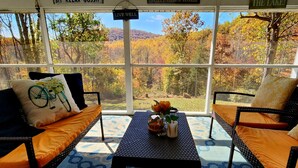 A covered outdoor seating area with wicker furniture, a patterned floor, and a view of autumn trees. - Hidden Gem on Hidden Valley Lane (Linden)