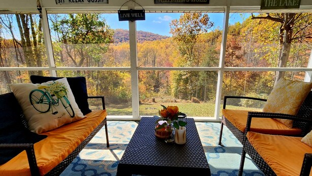 A covered outdoor seating area with wicker furniture, a patterned floor, and a view of autumn trees.