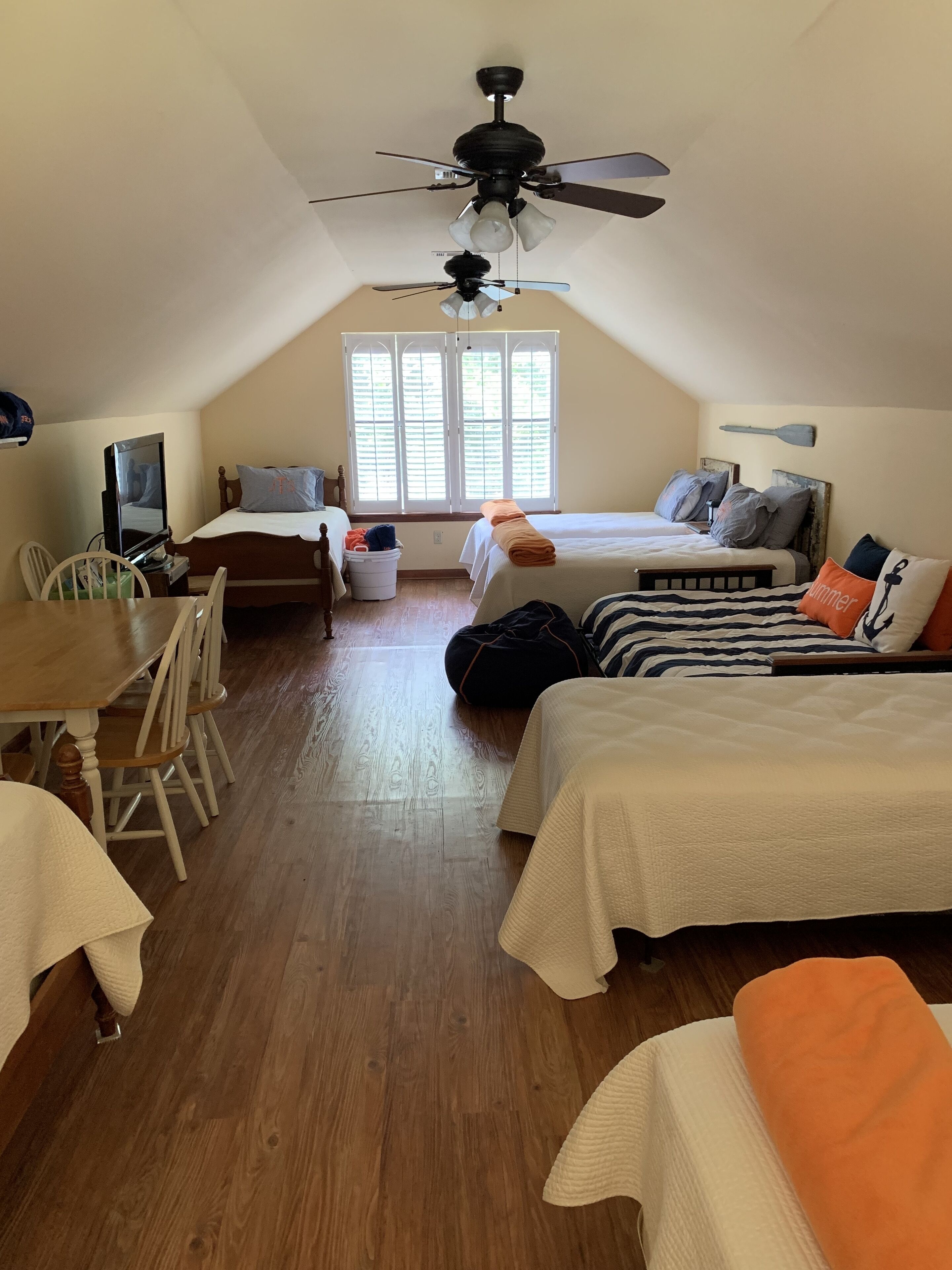 A cozy attic bedroom with a sloped ceiling, a ceiling fan, a large window with shutters, a bed with multiple pillows, a desk with a chair, and a television.