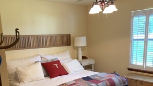 Un dormitorio con cama, ventilador de techo, mesita de noche, lámpara y ventana.