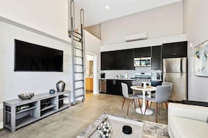 A modern living room with a flat-screen TV, a grey entertainment unit, and a white sofa. A kitchen with black cabinets and stainless steel appliances is visible in the background. - NEW Modern North Park. Short Walk From Everything (San Diego)
