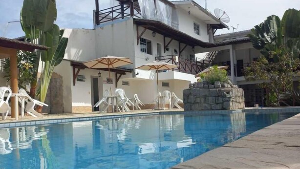 A swimming pool with lounge chairs and umbrellas, a white building with a balcony, and a stone structure. - Pousada Pérola do Mar (Tamandaré)