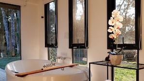 A modern bathroom with a freestanding bathtub, a wooden bath mat, and a black metal shelf with towels and a potted plant. - Reserva Cabanas (Balsa Nova)