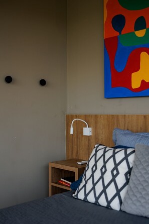Une chambre à coucher avec un lit, des oreillers, une table de chevet, une prise électrique murale et un tableau abstrait aux couleurs vives.