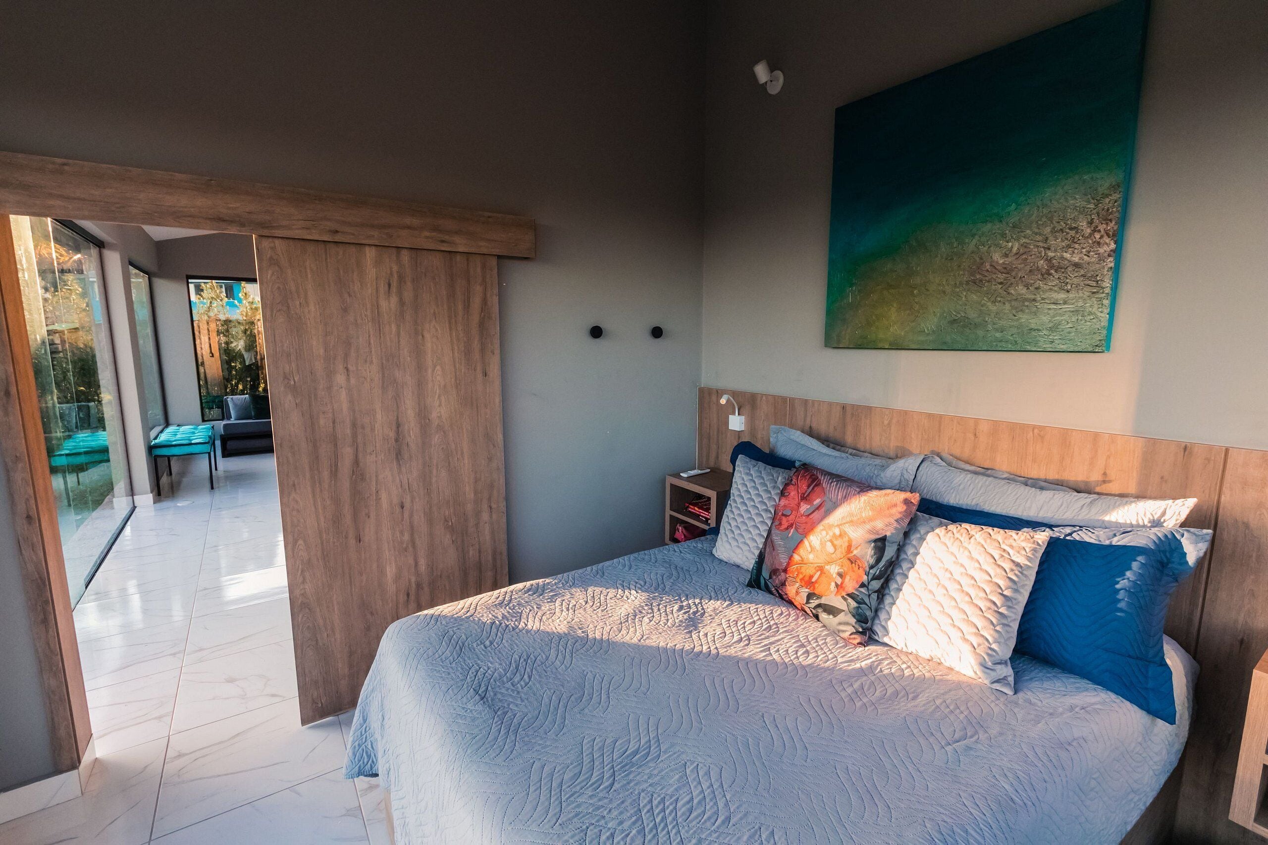 A bedroom with a large bed, a nightstand, a painting on the wall, and a view of the outside.