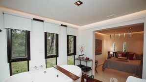A modern bathroom with a large bathtub, a small table with a glass of water, and a view of a bedroom with a bed and a nightstand. - Reserva Cabanas (Balsa Nova)