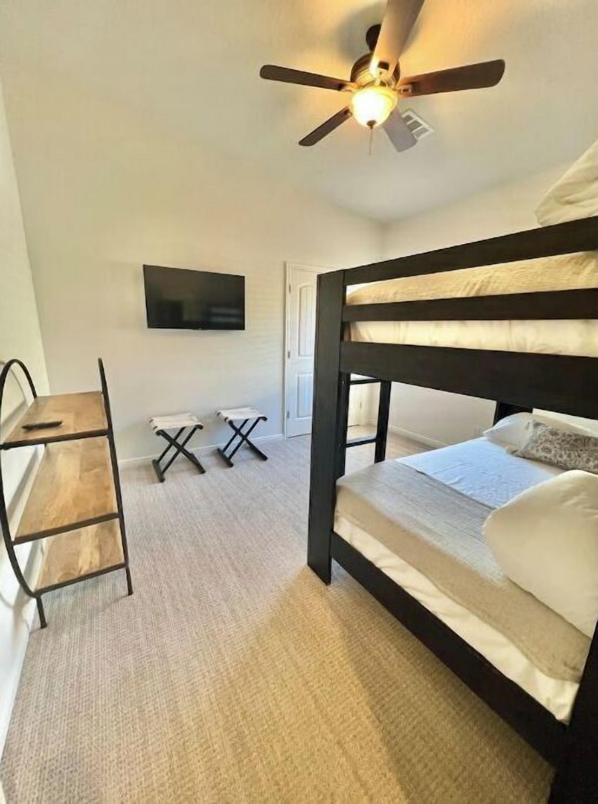 Une chambre avec un lit superposé, un bureau avec une chaise, un téléviseur fixé au mur et un ventilateur de plafond.