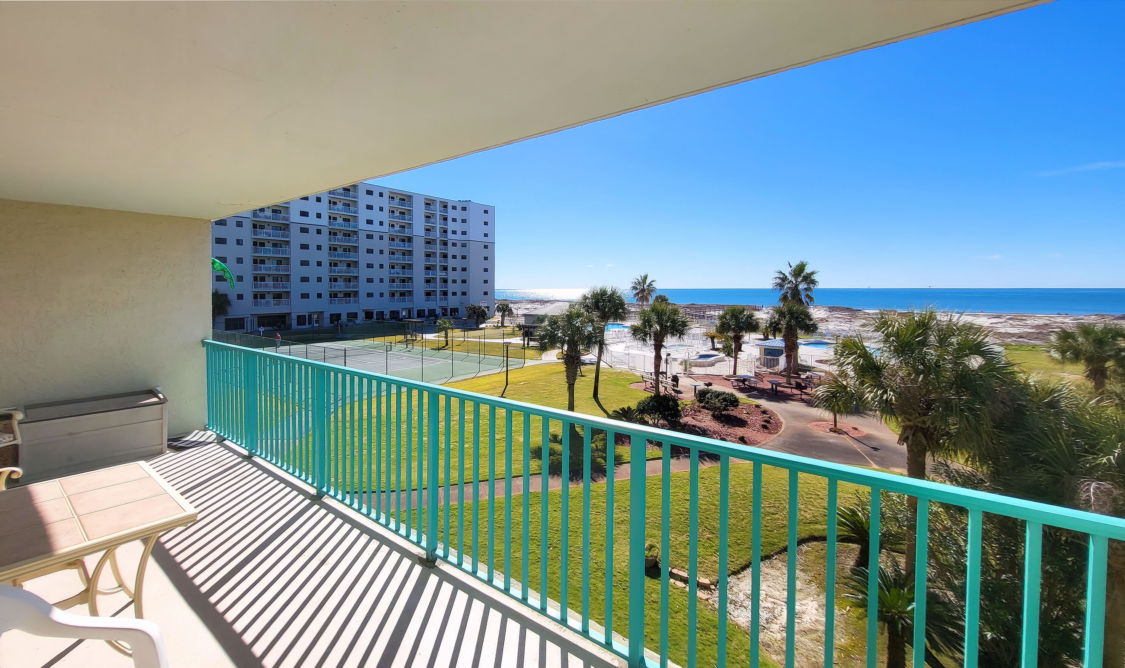 A balcony with a view of a beach, a tennis court, and a multi-story building.