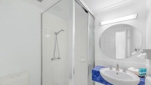 A bathroom with a glass shower enclosure, a white sink with a blue countertop, and a round mirror above the sink. - Amaroo at Trinity - Classic Ocean View Studio (Cairns)