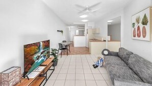 A modern living room with a sofa, a dining area with a table and chairs, a kitchen in the background, and a ceiling fan. - Amaroo at Trinity - Classic Ocean View Studio (Cairns)