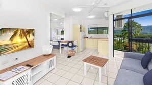 A modern living room with a sofa, a coffee table, a TV mounted on the wall, and a balcony with a view of palm trees and mountains. - Amaroo at Trinity - Classic Ocean View Studio (Cairns)