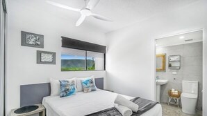Une chambre à coucher moderne, avec un lit, un ventilateur de plafond et une salle de bain visible à travers une porte ouverte.