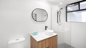A modern bathroom with a white toilet, a round mirror, a white sink with a wooden cabinet, and a glass shower enclosure. - Luxurious Studio with Lush Mountain Views (Cairns)