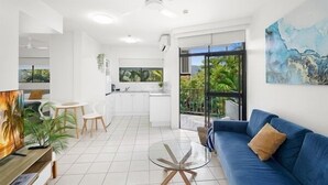 A modern living room with a blue sofa, a glass coffee table, and a TV. There is a kitchen area with white cabinets and a dining table with chairs. - Luxurious Studio with Lush Mountain Views (Cairns)