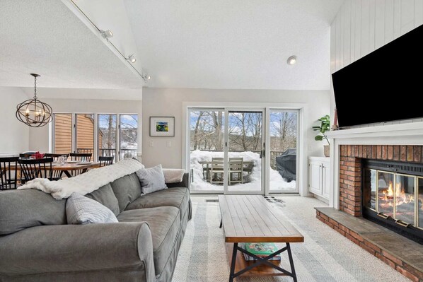 A modern living room with a fireplace, a dining area, and a view through glass doors. - End-Unit Condo with Sauna & Pool, Deere Run (Quechee)