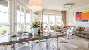 A modern living room with a glass dining table, clear chairs, a sofa, and a large window offering a view of the outdoors. - Alte Mühle, Whg. 12 Polo Suite - Alte Mühle (Sylt-Ost)