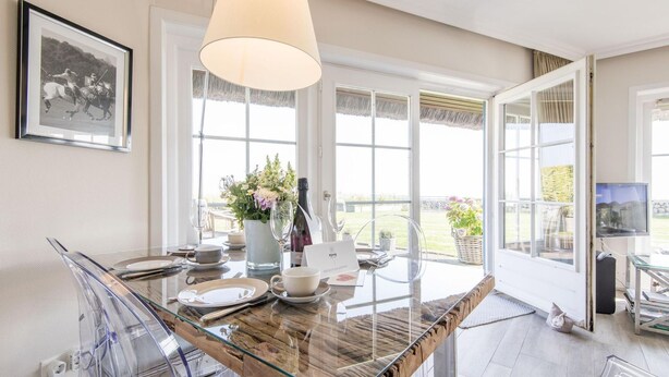 A modern kitchen with a glass dining table, clear chairs, a large window, and a framed picture on the wall. - Alte Mühle, Whg. 12 Polo Suite - Alte Mühle (Sylt-Ost)