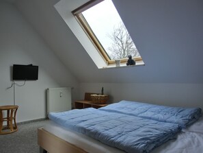 A bedroom with a sloped ceiling, a bed with blue bedding, a television mounted on the wall, a radiator, a small wooden stool, and a window with a view of trees.