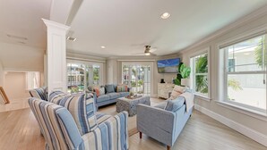 A spacious living room with a large sectional sofa, a gray armchair, and a coffee table. There are large windows providing natural light and a view of the outdoors.