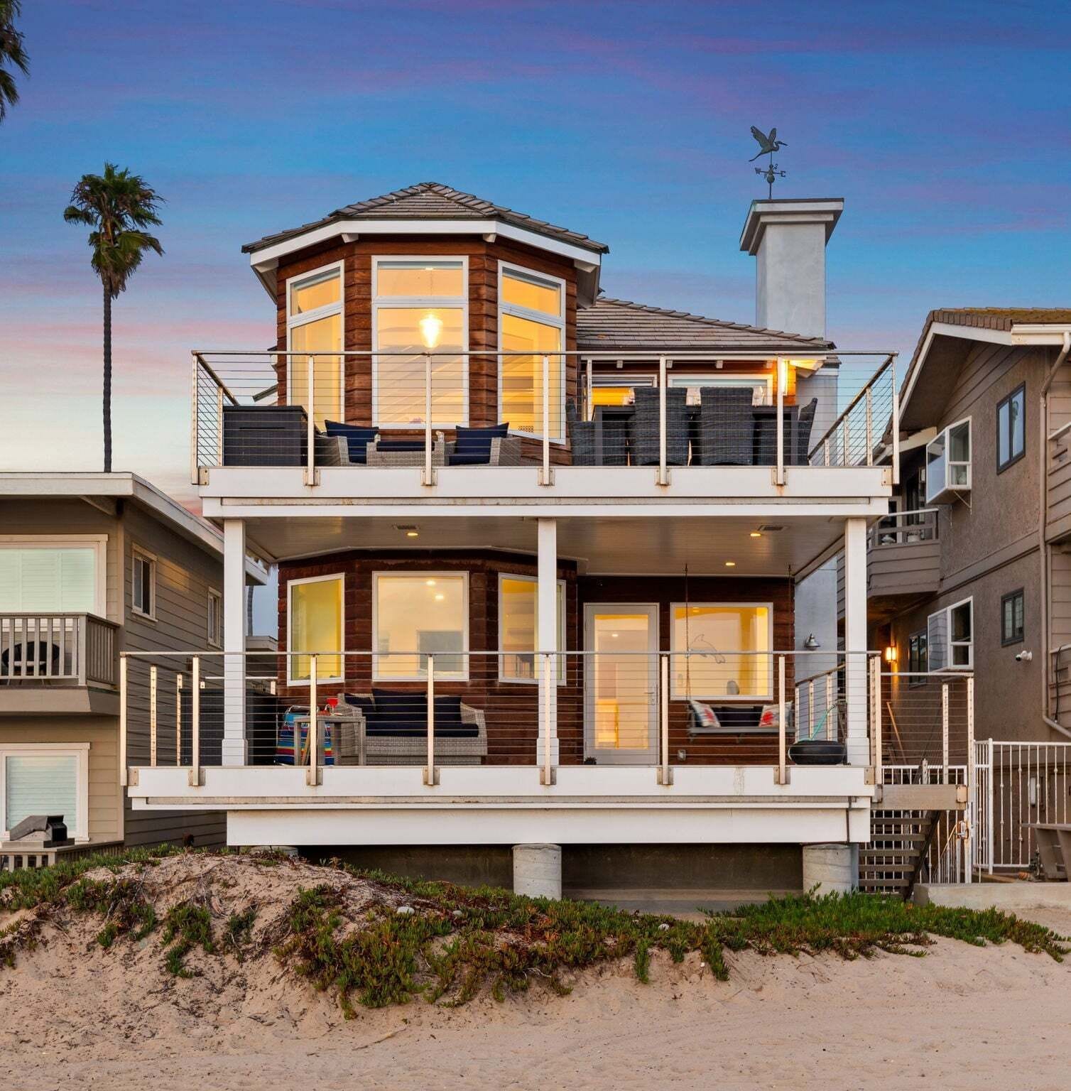 A two-story beachfront house with a balcony, large windows, and a weather vane.