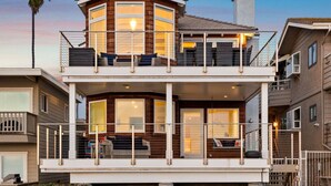 A two-story beachfront house with a balcony, large windows, and a weather vane.