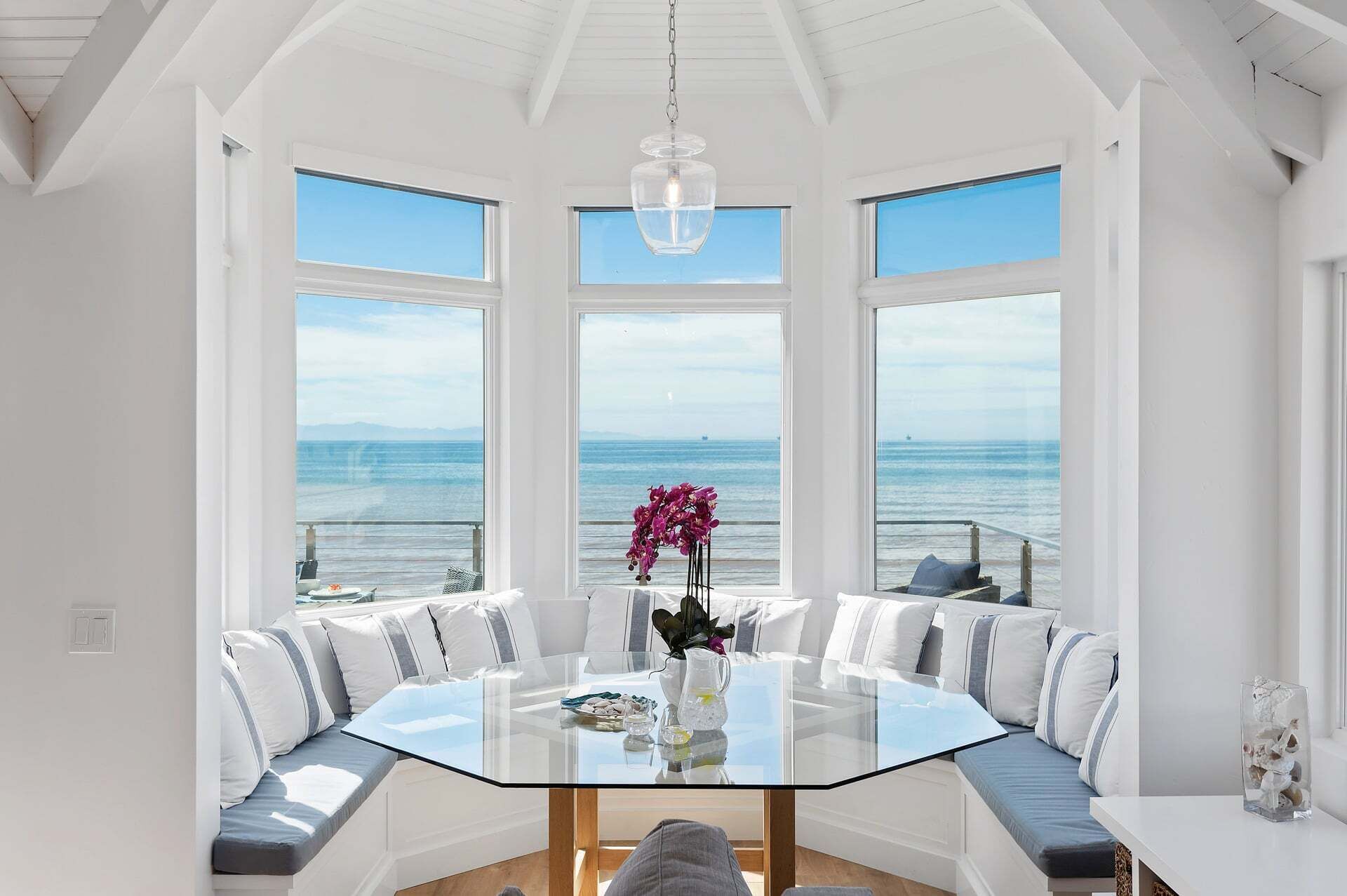 A spacious dining area with a glass table and white upholstered seating, large windows with ocean views, and a hanging light fixture.