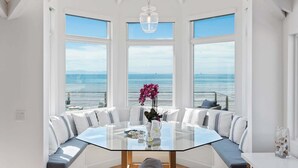 A spacious dining area with a glass table and white upholstered seating, large windows with ocean views, and a hanging light fixture.