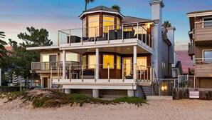 A two-story beachfront house with a balcony, a covered patio, and a view of the ocean.