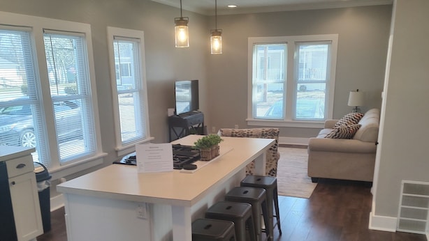 Una cocina moderna con una isla central, taburetes de bar e iluminación colgante. Hay un área de comedor con un sofá y una lámpara.