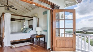 A bedroom with a wooden ceiling, a bed with white bedding, a nightstand, and a view of the landscape through a large window.