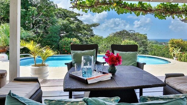 A covered outdoor seating area with wicker furniture, a table with drinks and a book, overlooking a pool and lush greenery.