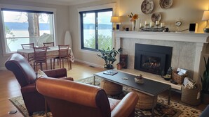 A living room with a fireplace, a dining area with a table and chairs, and a view of the outdoors. - Amazing Views and Whale Watching from Indoors and Outside, with AC! (Camano)
