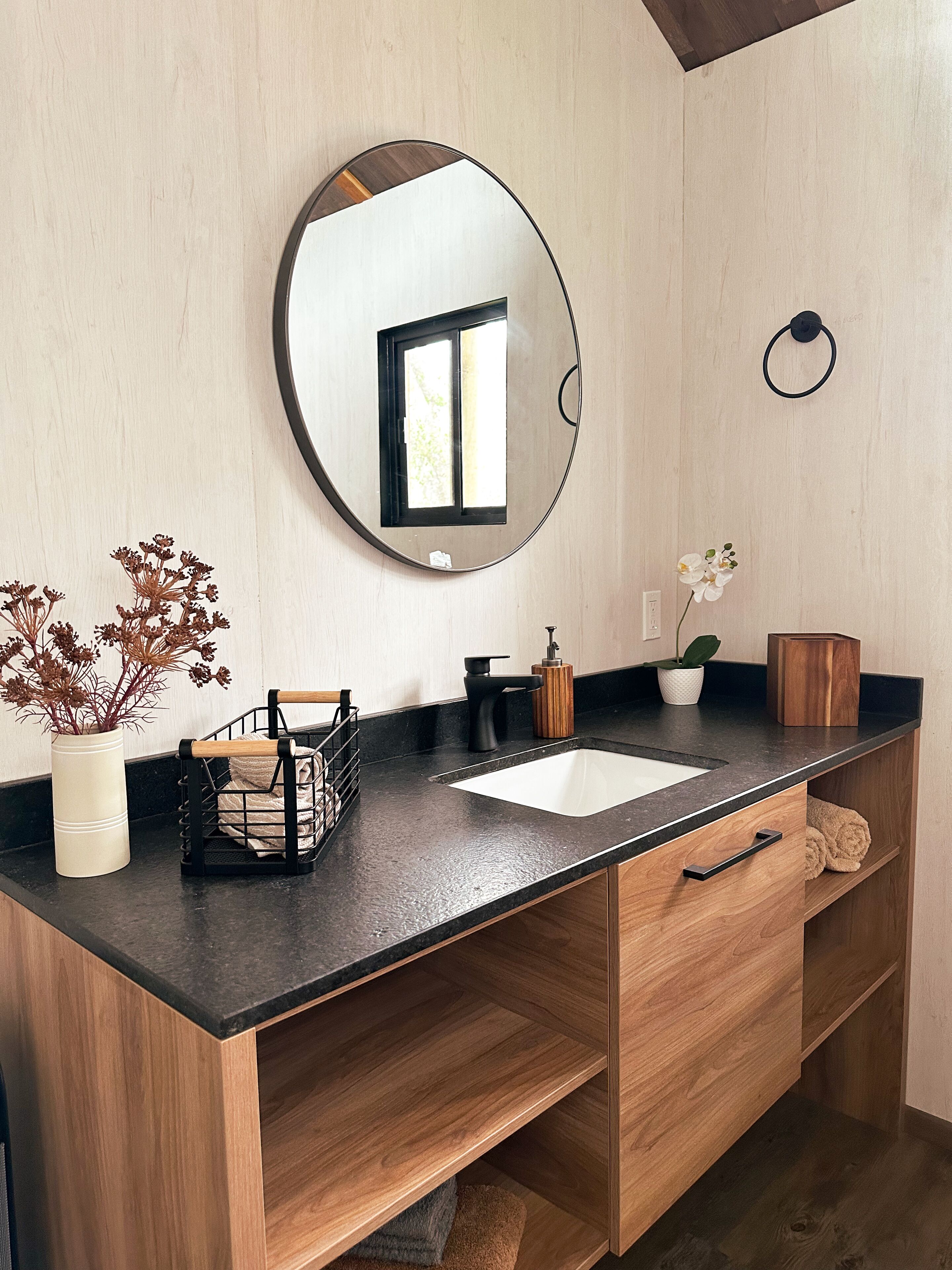 A modern bathroom with a black countertop, a white sink, a wooden vanity, a round mirror, a small window, and a potted plant.