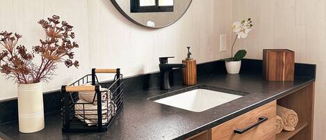 A modern bathroom with a black countertop, a white sink, a wooden vanity, a round mirror, a small window, and a potted plant.