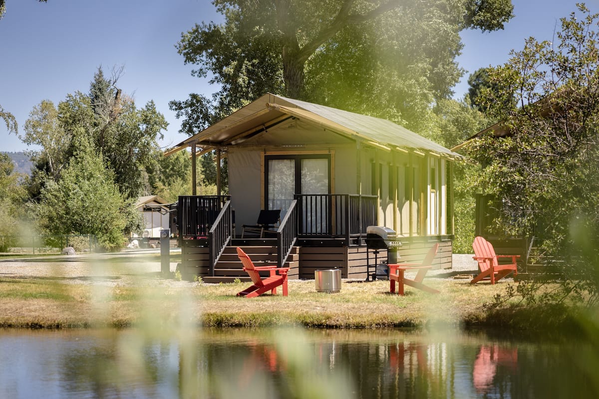 A small cabin with a porch, outdoor seating, and a pond in front.