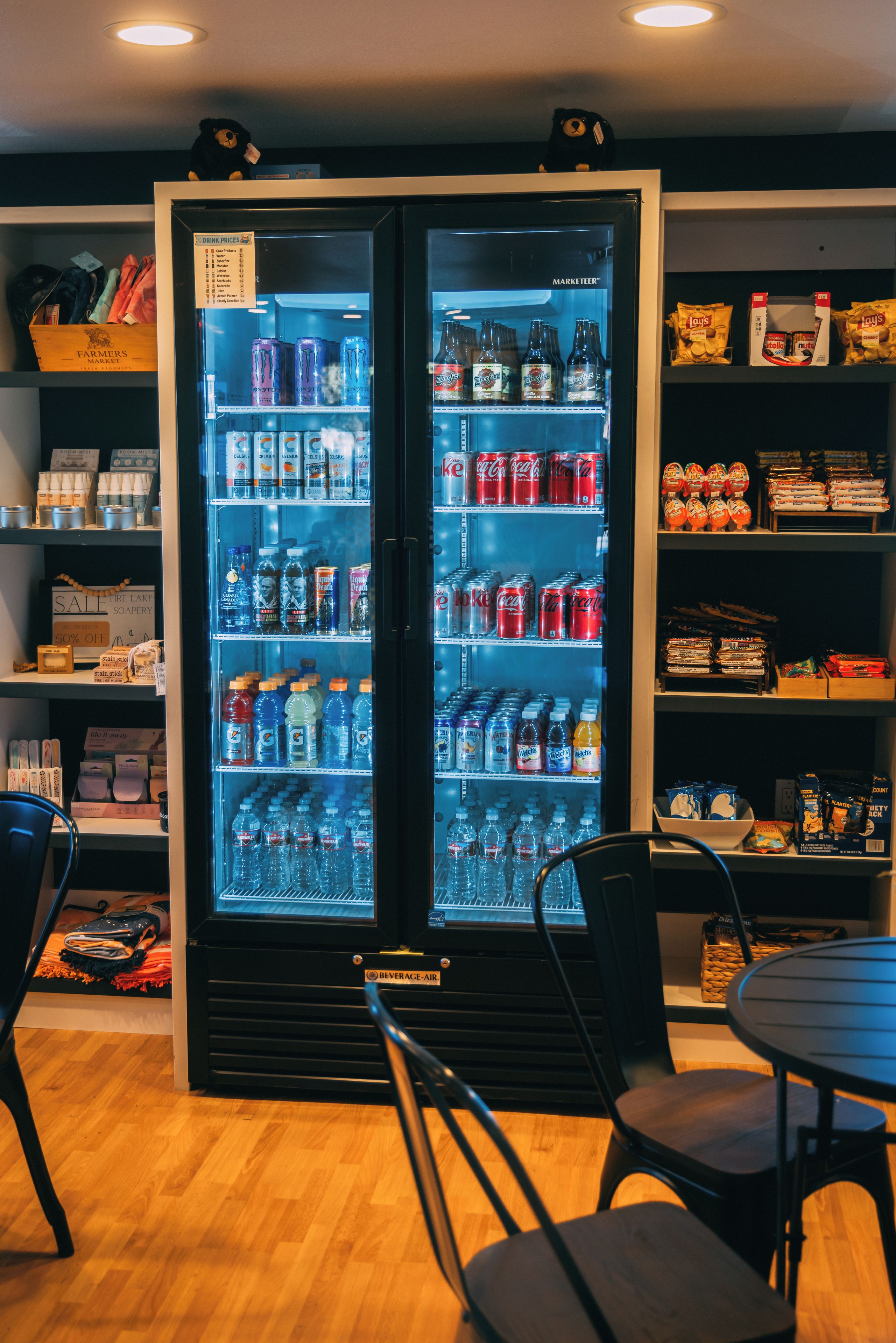 Un réfrigérateur rempli de diverses boissons, une boîte en bois étiquetée « Marché de producteurs » et des étagères contenant des collations et d’autres articles.