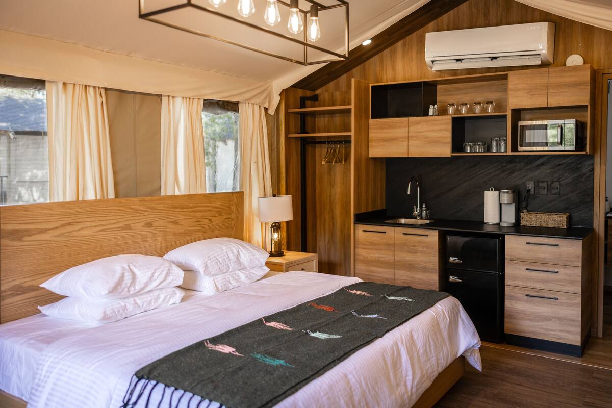 A bedroom with a bed, bedside table, lamp, and a kitchenette area with a microwave and cabinets.