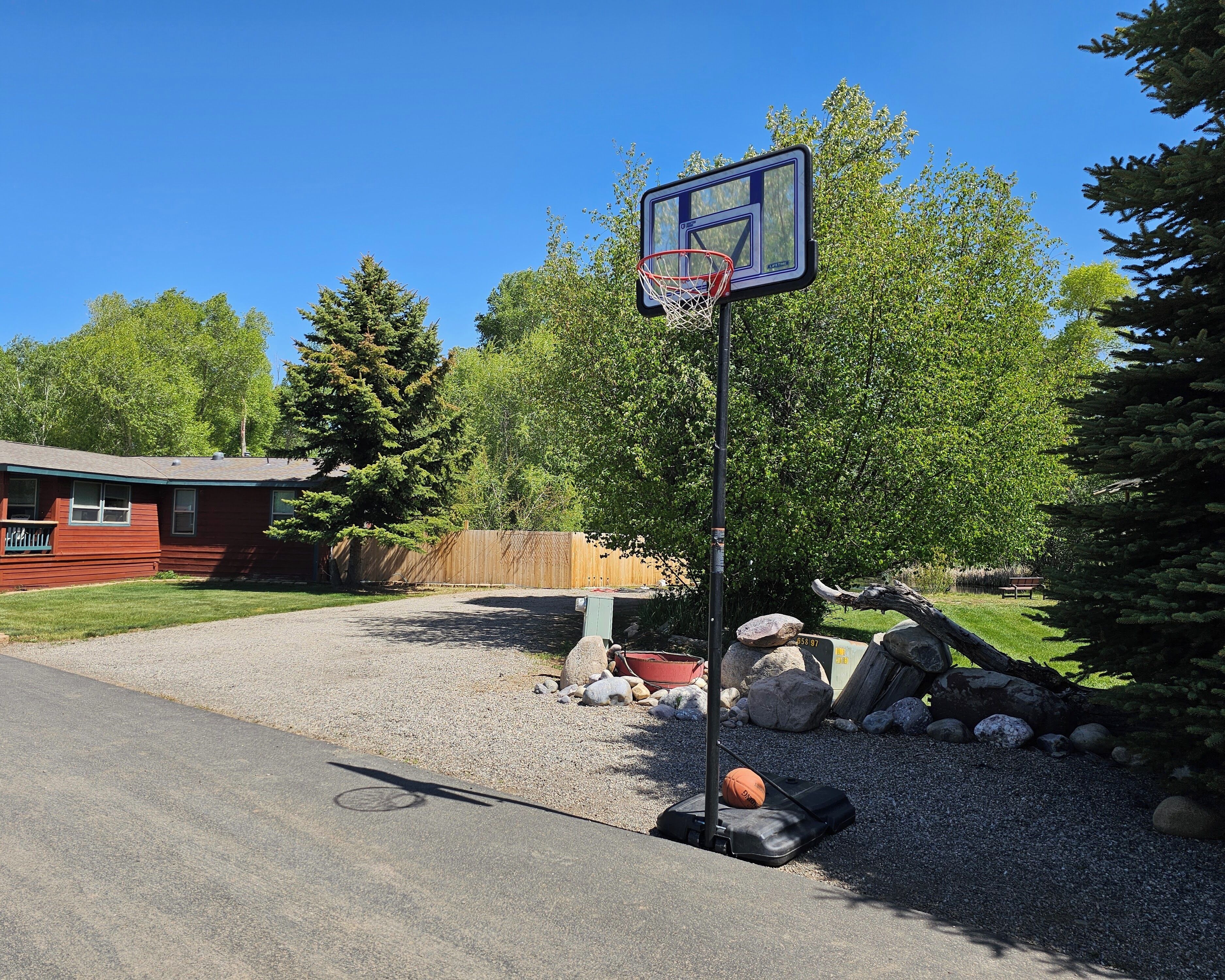 A basketball hoop in a residential area with a gravel driveway, a wooden fence, and a red building in the background.