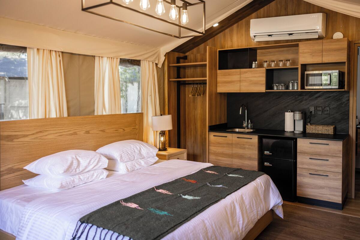 A bedroom with a bed, bedside table, lamp, and a kitchenette area with a microwave and cabinets.