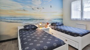 A bedroom with two beds, a mural of a beach scene, a window with curtains, and a nightstand with a lamp.