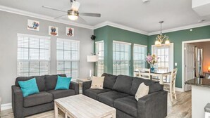 Ein Wohnzimmer mit zwei dunkelgrauen Sofas, einem Holztisch und einem Deckenventilator. Große Fenster mit Jalousien, ein Essbereich mit Tisch und Stühlen sowie eine Küchenzeile mit Arbeitsplatte und Spüle.