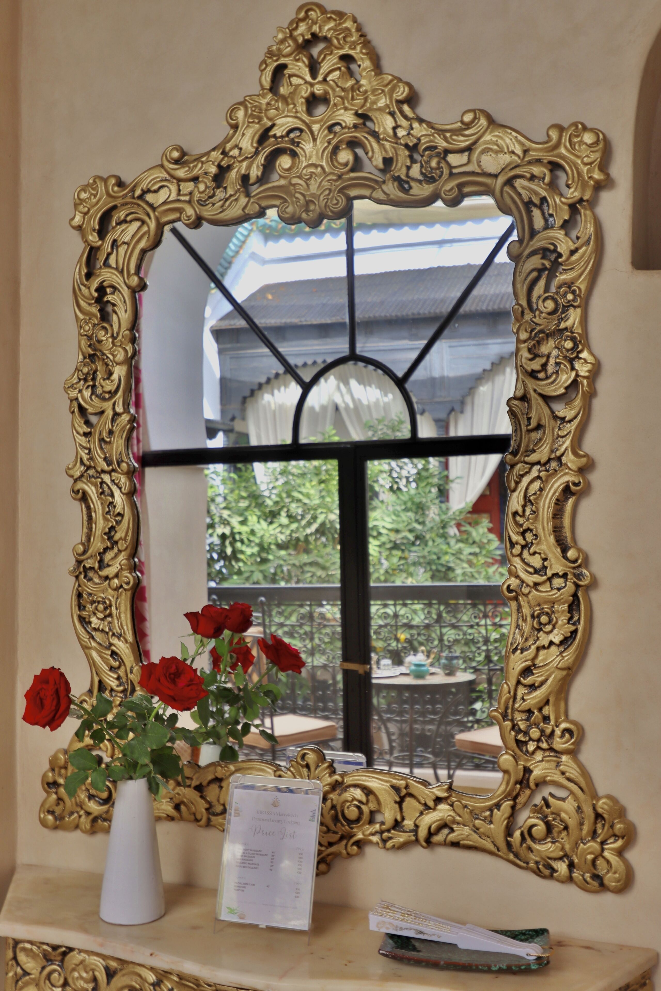 Un miroir à cadre doré, avec un vase blanc contenant des roses rouges et un menu posé sur une surface en marbre.