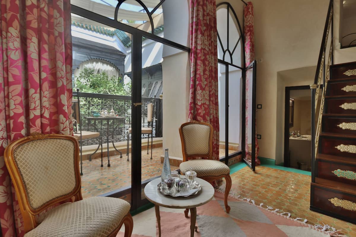 A room with two wicker chairs, a small round table with a tray of items, a view of a garden through a glass door, and a staircase with ornate railings.