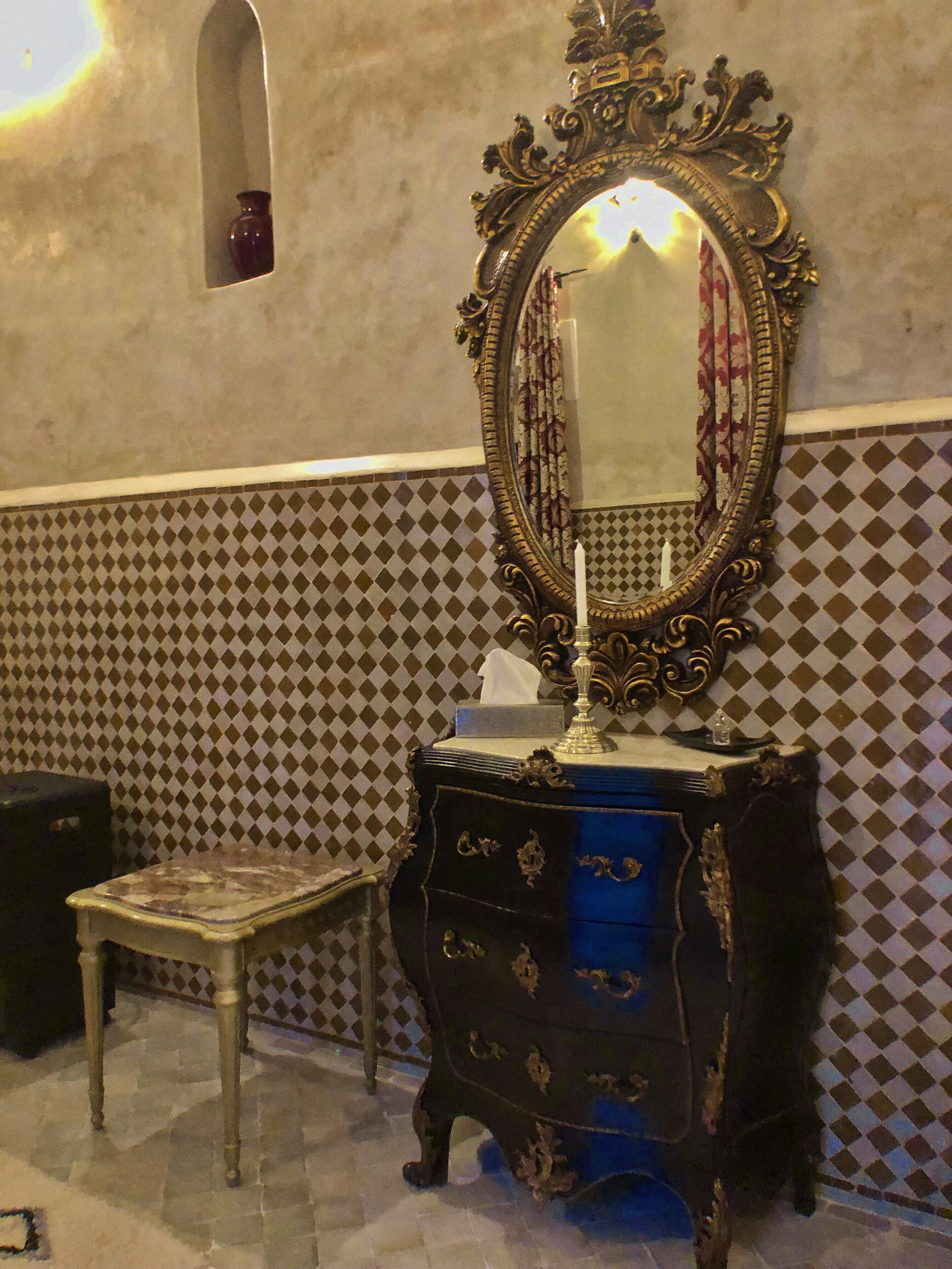 A bathroom with a decorative mirror, a vanity with drawers, a small stool, and a patterned wall.