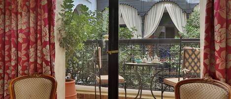 A room with two wicker chairs, a small table with a tray of items, and a view of an outdoor balcony with a tiled floor and potted plants.